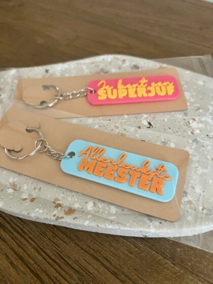 3D-geprinte sleutelhangers 'Je bent een Superjuf' in roze en 'Allerleukste Meester' in lichtblauw, verpakt op kraftkaartjes